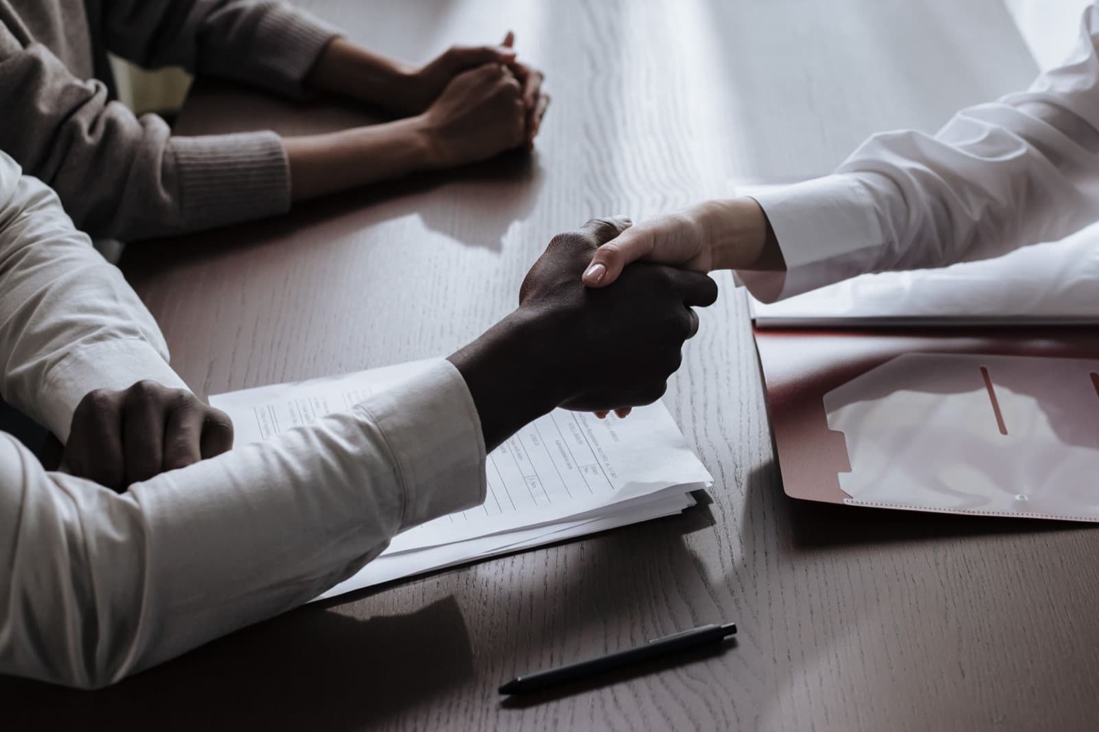 Two people shaking hands across a meeting table over contract documents and a folder — closing a negotiated elevator contract