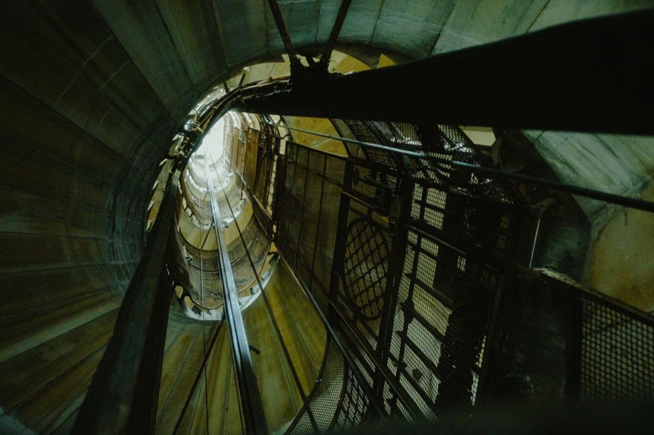 Looking up an elevator shaft at guide rails, cables, and hoisting equipment — the components behind emergency elevator repairs