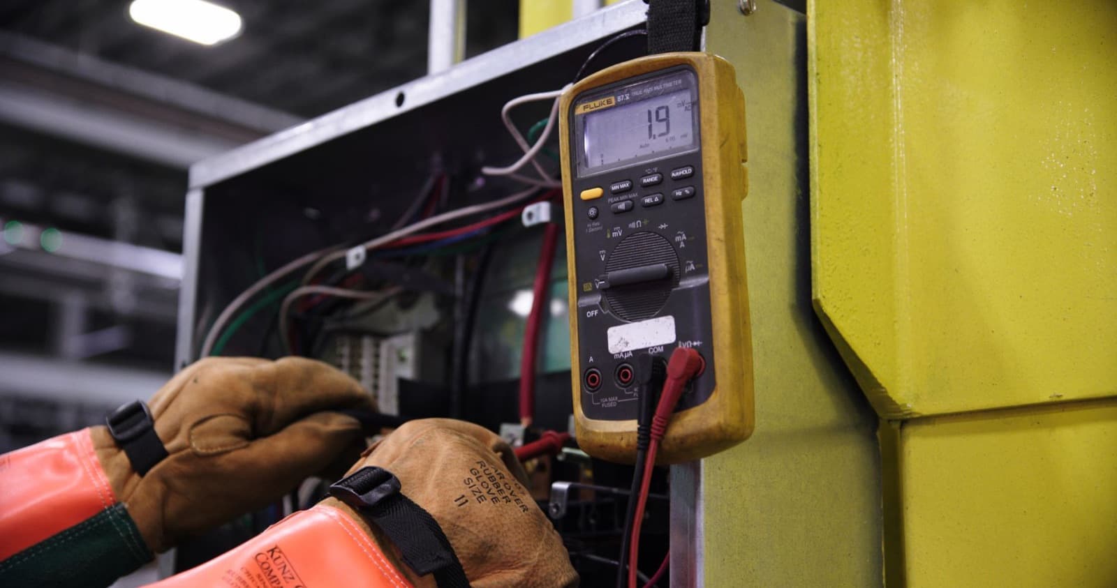 Technician in insulated gloves testing an elevator control cabinet with a multimeter — what drives elevator repair cost